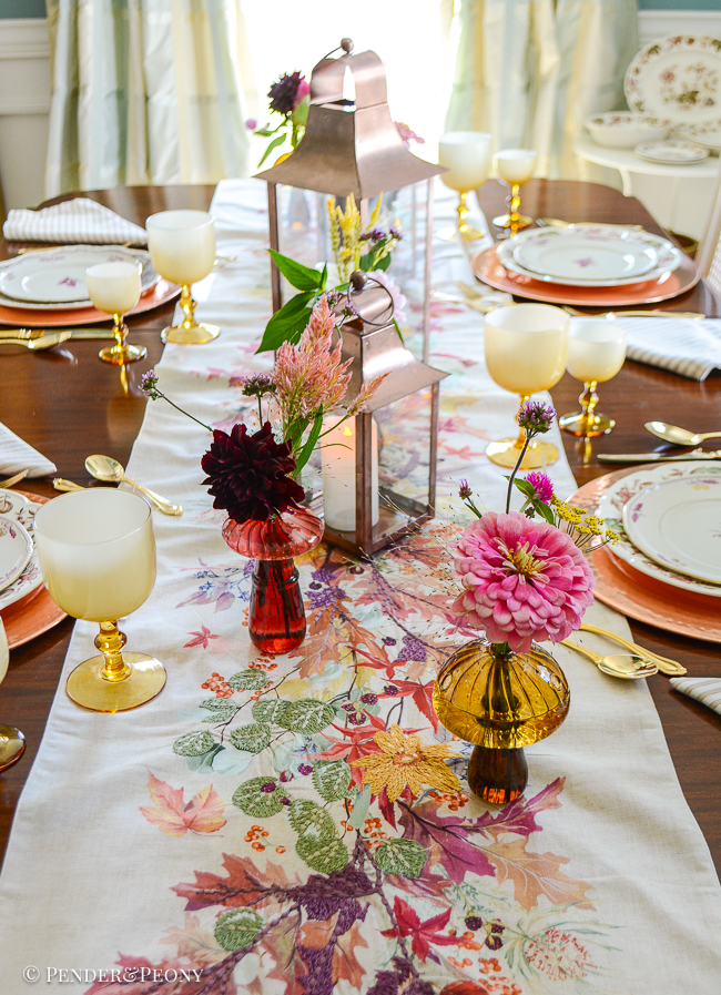 An embroidered floral runner decorates the center of this autumn tablescape with glass mushroom bud vases, wildflowers, and copper lanterns.
