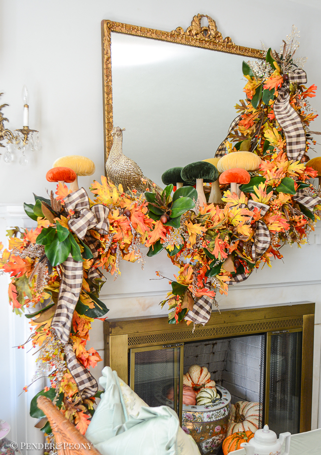 A fall mantel garland created with orange and gold oak leaves, bittersweet, magnolia, glittered fern, brown and white gingham ribbon. Mantel top decorated with velvet mushrooms and pheasant.