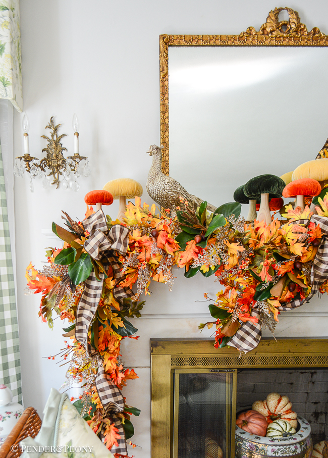 A fall mantel garland created with orange and gold oak leaves, bittersweet, magnolia, glittered fern, brown and white gingham ribbon. Mantel top decorated with velvet mushrooms and pheasant.