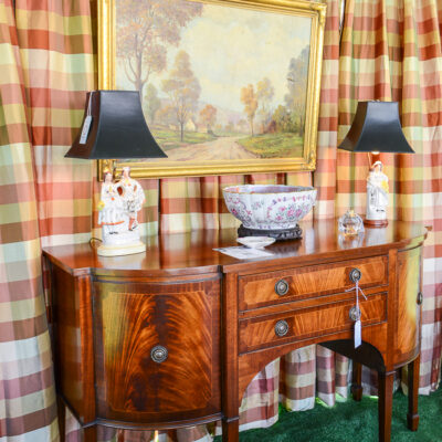Back wall of design tent hung with silk plaid curtains in pink, burgundy, and green with English mahogany sideboard, landscape painting, and Staffordshire flatback lamps