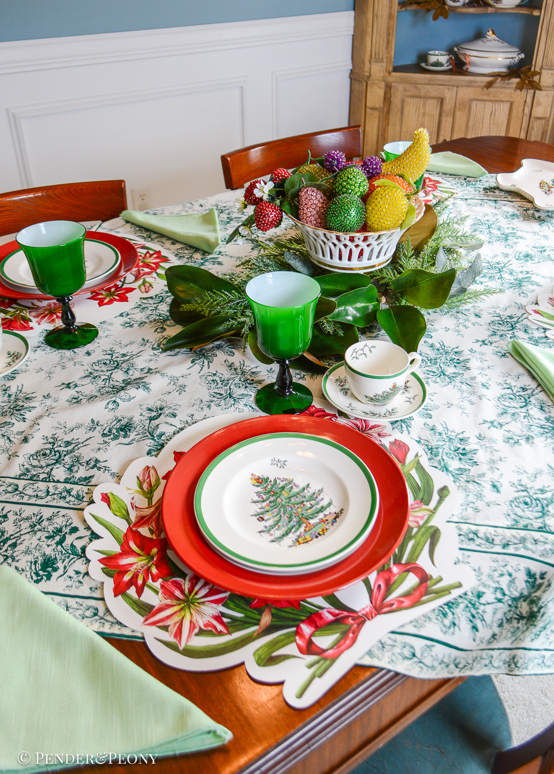 Green, white, and red holiday tablescape with Spode's Christmas tree dishes, amaryllis placemats, vintage beaded fruit, and Moretti emerald goblets