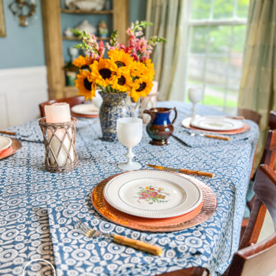 A blue and white summer table with Wedgwood dishes, copper chargers, white Portieux Vallerysthal goblets, and sunflowers in a blue spongeware jug