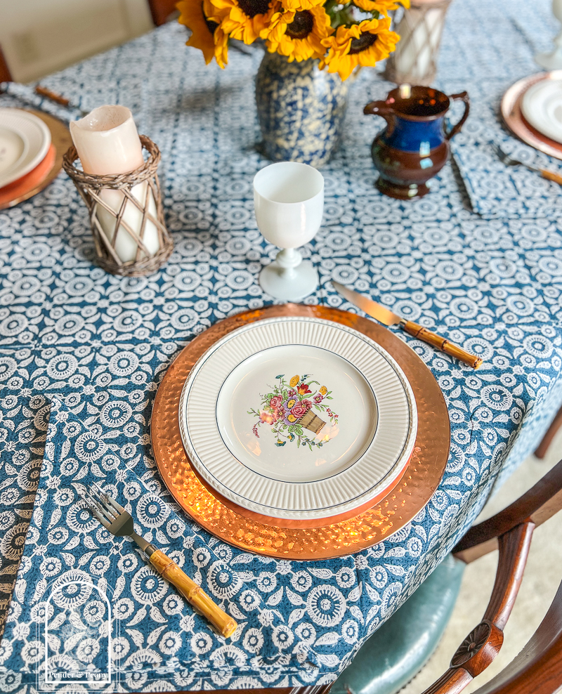 A blue and white summer table with Wedgwood dishes, copper chargers, white Portieux Vallerysthal goblets, and sunflowers in a blue spongeware jug
