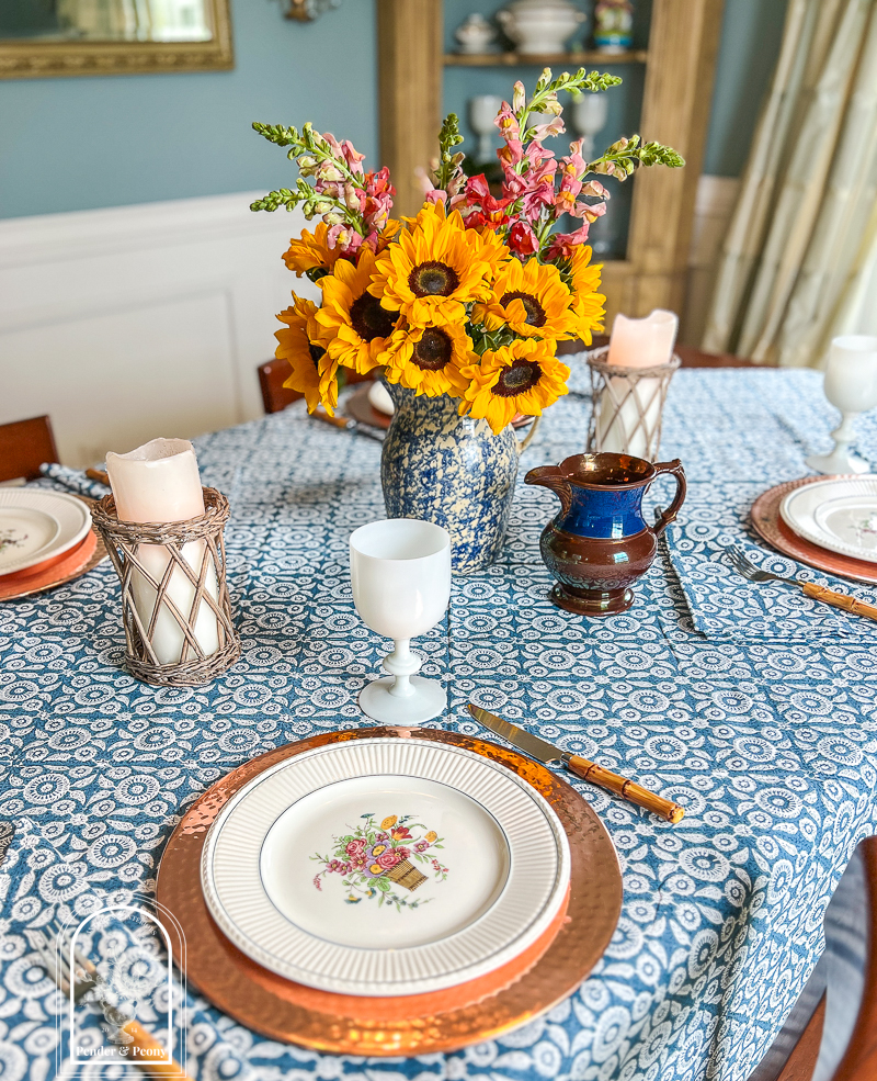 A blue and white summer table with Wedgwood dishes, copper chargers, white Portieux Vallerysthal goblets, and sunflowers in a blue spongeware jug