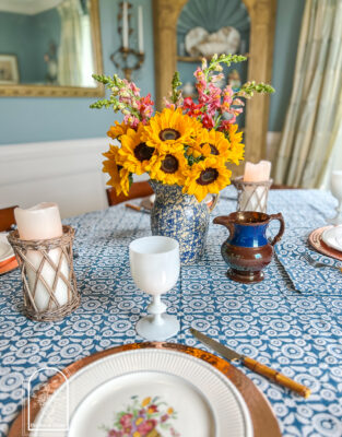 A blue and white summer table with Wedgwood dishes, copper chargers, white Portieux Vallerysthal goblets, and sunflowers in a blue spongeware jug