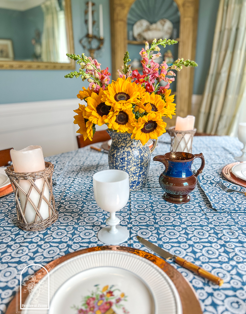 A blue and white summer table with Wedgwood dishes, copper chargers, white Portieux Vallerysthal goblets, and sunflowers in a blue spongeware jug