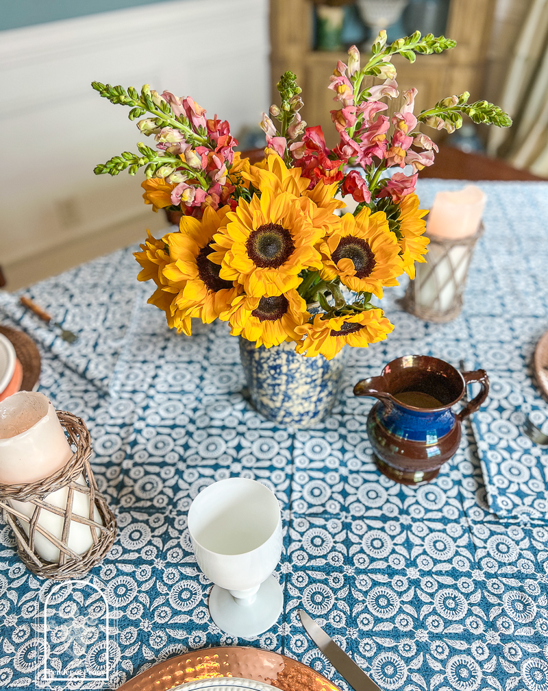 A blue and white summer table with Wedgwood dishes, copper chargers, white Portieux Vallerysthal goblets, and sunflowers in a blue spongeware jug