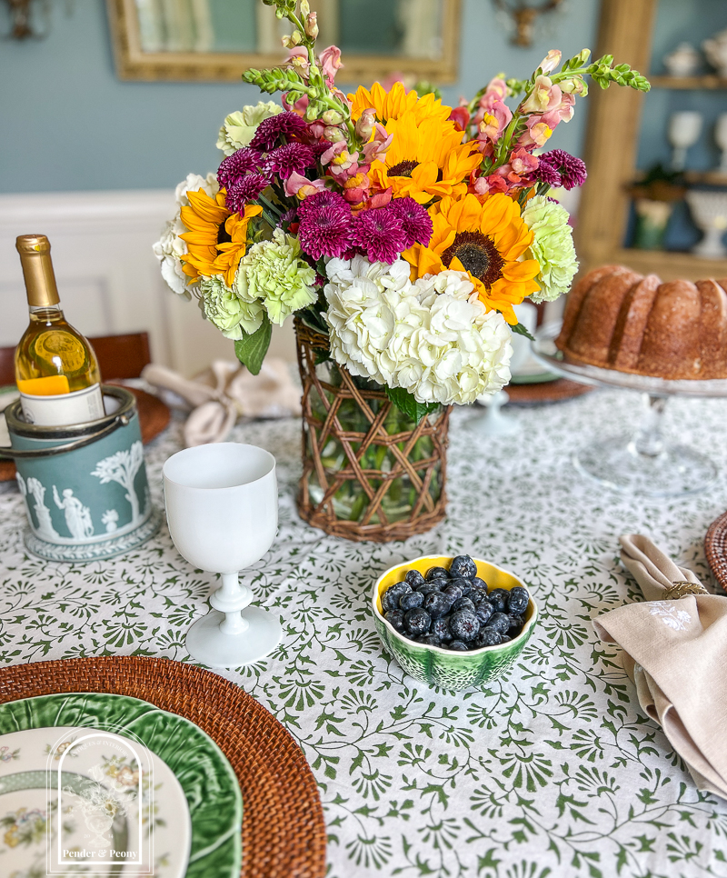 A summer floral centerpiece with hydrangea, sunflowers, snap dragons, and mums in a wicker wrapped vase.
