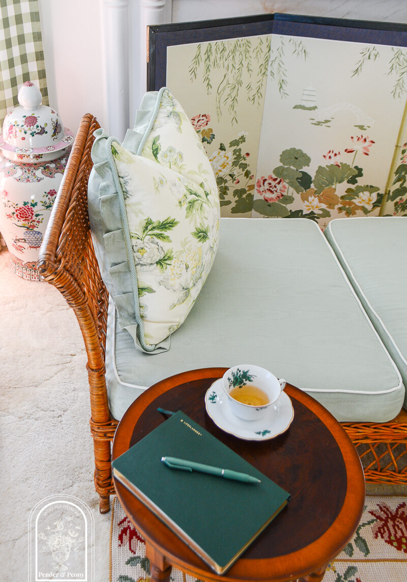 Wicker daybed with chintz pillow and side table with china teacup and green leather note book - cultivate beauty in your home with just a few small efforts