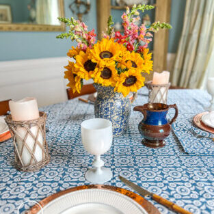 A blue and white summer table with Wedgwood dishes, copper chargers, white Portieux Vallerysthal goblets, and sunflowers in a blue spongeware jug