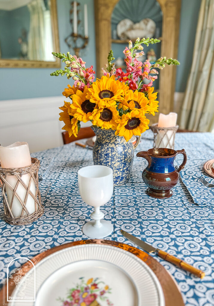 A blue and white summer table with Wedgwood dishes, copper chargers, white Portieux Vallerysthal goblets, and sunflowers in a blue spongeware jug