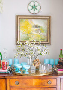 Mahogany sideboard styled as bar with aqua opaline glasses, dogwood branch, and brass tray