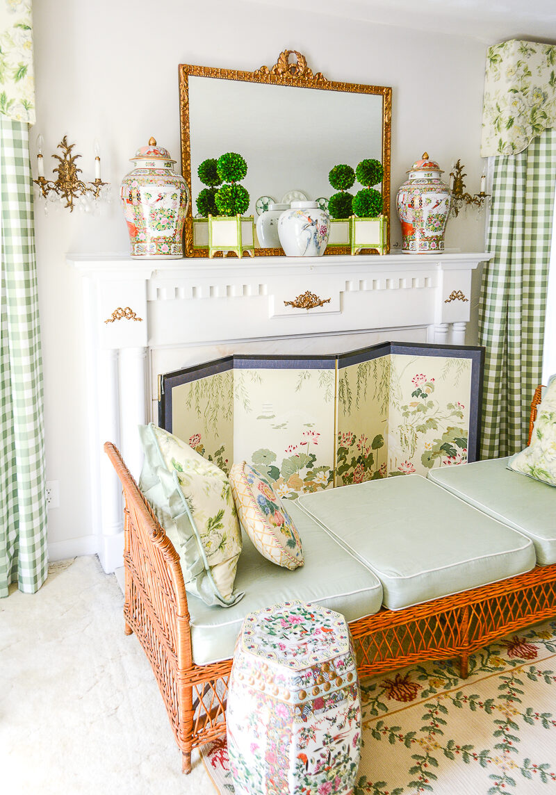 My Grandmillennial living room mantel with boxwood topiary, a gilt French mirror, Rose Medallion temple jars, and Chinese silk screen. A wicker day bed sits in front with garden stools. All decor and furnishings were collected second hand at auctions, estate sales, and thrifts.